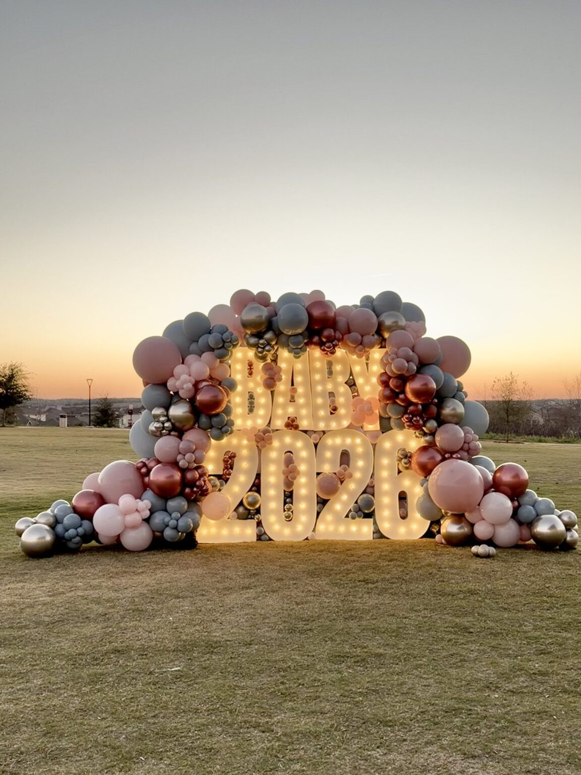 Dusty rose and burgundy balloon garland around glowing BABY 2026 marquee letters on a lawn at sunset in Austin, TX