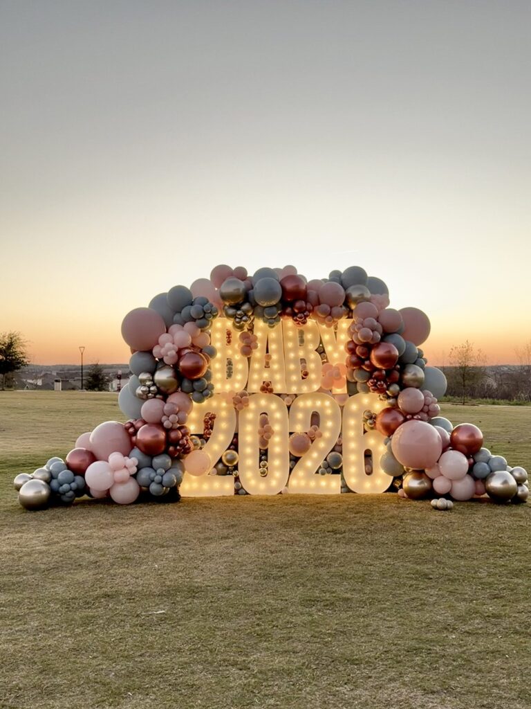 Dusty rose and burgundy balloon garland around glowing BABY 2026 marquee letters at sunset in Austin, TX