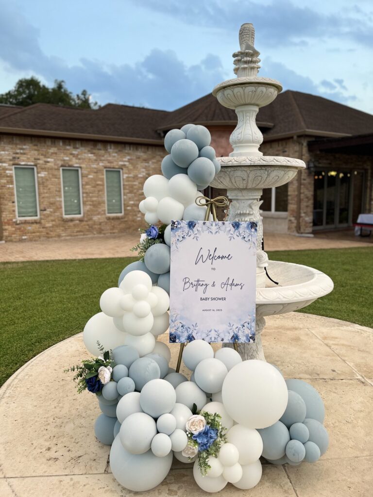 Dusty blue and white balloon garland climbing a stone fountain at an outdoor baby shower in Austin, TX