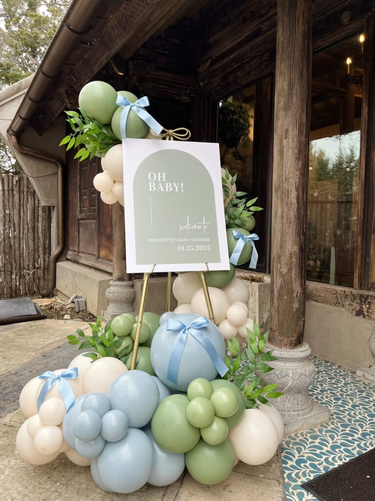 Sage green and dusty blue balloon garland with satin bows around an Oh Baby shower sign in Austin, TX